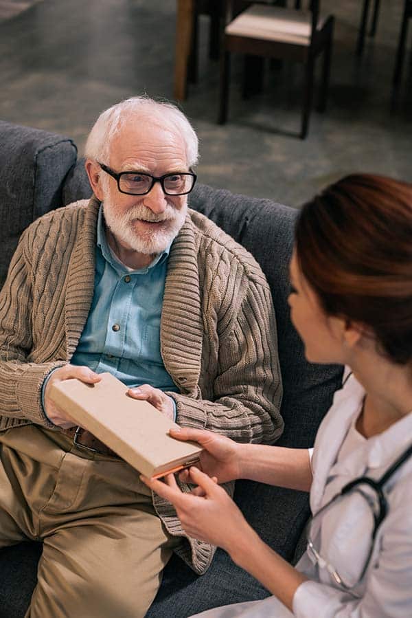 Female nurse handing a book to an older man on a couch